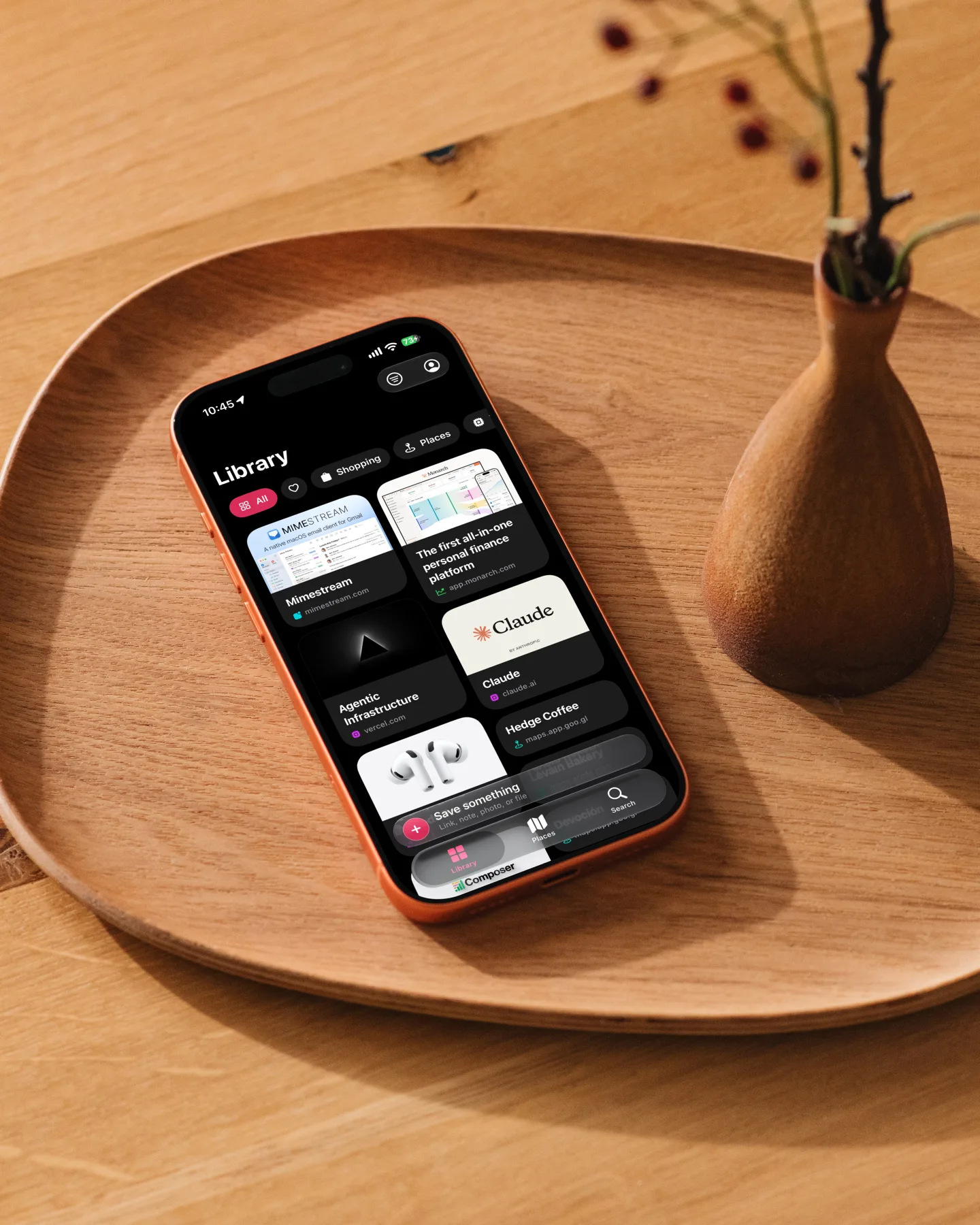 An iPhone lying on a wooden serving tray beside a small wooden vase, showing the FaveNest Library in dark mode: a grid of saved cards including a Mac email app, a personal-finance site, Claude, and a café saved from Google Maps.